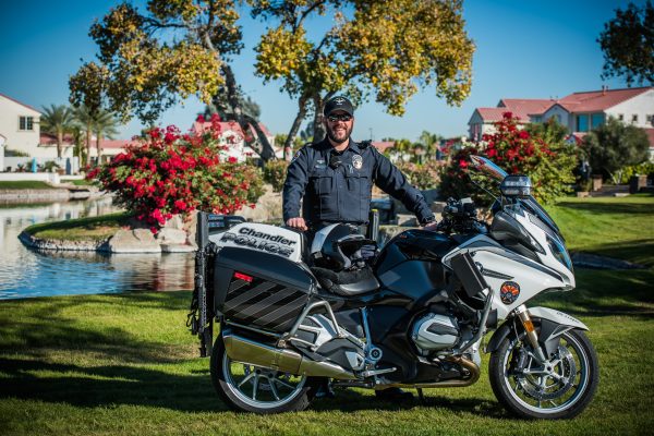 Chandler PD Motor officer standing behind his motorcycle with a housing area pond and garden landscape in the background.